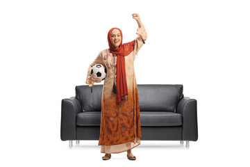 Wall Mural - Young woman in ethnic clothes holding a football and cheering in front of a sofa