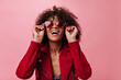 © Look! - Joyful girl in red jacket takes off sunglasses on isolated background. Woman in good mood smiling on pink backdrop