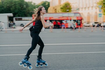 Wall Mural - Outdoor shot of active slim young woman enjoys rollerskating during spare time dressed in blackactive wear poses in urban place on road against blurred background with transport. Hobby concept