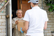 © Angelov - Young delivery man in uniform giving a box, parcel of groceries to elderly man outdoor. Shopping help and delivery service. Volunteer support seniors during coronavirus outbreak