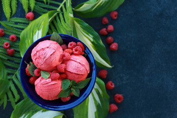 Summer dessert. Raspberry ice cream balls in blue bowl with fresh berries: raspberries and strawberries with mint and herbs, leaves on black background. Tropical concept. Background image, copy space