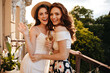 © Look! - Portrait of two charming girls smiling on terrace. Young red-haired woman with her friend in straw hat posing at camera with glass of champagne