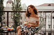 © Look! - Charming woman in floral outfit holds wine glass and rests on terrace. Red-haired girl with champagne sits on white modern chair and relaxes.