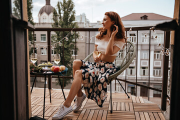 Charming woman in floral outfit holds wine glass and rests on terrace. Red-haired girl with champagne sits on white modern chair and relaxes.