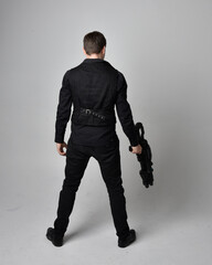 Wall Mural - Full length portrait of a  brunette man wearing leather jacket  and holding a science fiction gun.  Standing  action pose isolated  against a grey studio background.