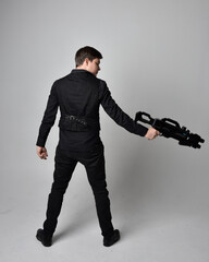 Full length portrait of a  brunette man wearing leather jacket  and holding a science fiction gun.  Standing  action pose isolated  against a grey studio background.