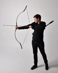 Wall Mural - Full length portrait of a  brunette man wearing black shirt and waistcoat holding  a bow and arrow.  Standing  action pose isolated  against a grey studio background.