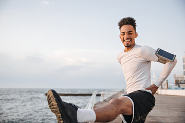 Wall Mural - Happy excited brunette dark-skinned bearded man in white t-shirt and black shorts stretches and smiles near sea.