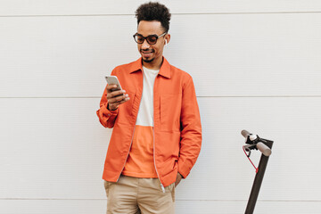 Wall Mural - Handsome curly brunet man in sunglasses, orange jacket and beige shorts smiles sincerely, holds phone and poses near scooter and white wall.