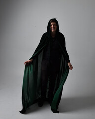 Full length portrait of a  brunette man wearing black shirt, waistcoat and a green velvet cloak.  Standing  action pose isolated  against a grey studio background.