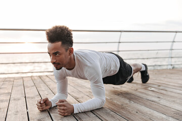 Wall Mural - Strong self-confident curly dark-skinned man in black shorts and white t-shirt does plank and works out near sea.