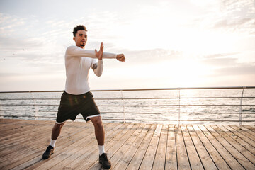 Wall Mural - Attractive charming dark-skinned sportsman in black shorts and white long-sleeved t-shirt does ark exercises, stretches and works out near sea.