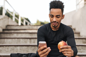 Wall Mural - Cool young brunette man in black long-sleeved t-shirt looks into phone screen, sits on stairs outside and holds fresh apple.