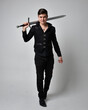 Full length portrait of a  brunette man wearing black shirt and gothic waistcoat holding sword.  Standing  action pose isolated  against a grey studio background.
