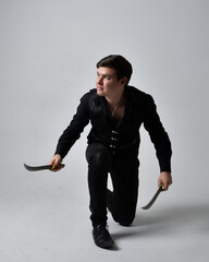 Wall Mural - Full length portrait of a  brunette man wearing black shirt and gothic waistcoat holding sword.  Standing  action pose isolated  against a grey studio background.
