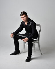 Wall Mural - Full length portrait of a  brunette man wearing black shirt and gothic waistcoat holding sword.  Seated  pose isolated  against a grey studio background.