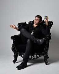 Full length portrait of a  brunette man wearing black shirt and  waistcoat.  Seated  pose in a gothic throne chair isolated  against a grey studio background.