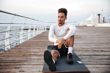 Wall Mural - Serious young dark-skinned man in white long-sleeved t-shirt and black shorts sits on yoga mat and listens to music in headphones near sea.