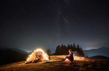 young man traveler sitting inside illuminated camp tent and playing guitar while spending time with 