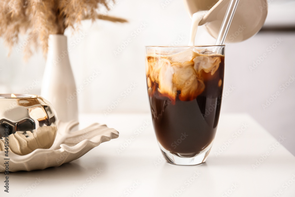 Pouring milk from jug into glass with tasty ice coffee on table, closeup - obrazy, fototapety, plakaty 
