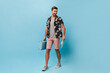 © Look! - Full length photo og stylish man in painted shirt, grey t-shirt, shorts and modern sneakers looking away, smiling and holding blue suitcase..