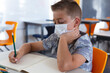 © WavebreakMediaMicro - Caucasian schoolboy wearing face mask sitting at desk in classroom writing in book