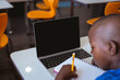 African american schoolboy sitting in classroom writing and using laptop, with copy space on screen