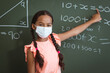 © WavebreakMediaMicro - Portrait of mixed race schoolgirl in face mask standing in front of chalkboard pointing in classroom