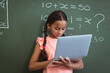 Mixed race schoolgirl standing in front of chalkboard in classroom using laptop