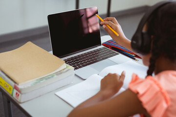 Wall Mural - Mixed race schoolgirl in classroom wearing headphones and using laptop, with copy space on screen