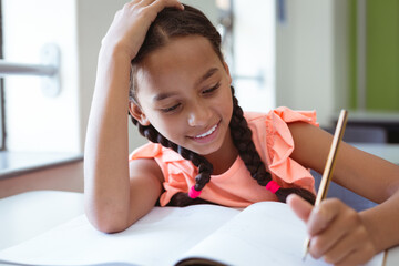 Wall Mural - Happy mixed race schoolgirl in classroom sitting at desk smiling and writing in book