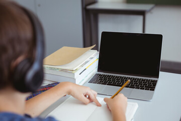 Wall Mural - Caucasian schoolboy in classroom wearing headphones and using laptop, with copy space on screen