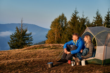 Wall Mural - Side view of married couple of travelers resting in tent on mountain meadow during autumn hike against the backdrop of trees and mountain hill. Sunshine morning in campsite.