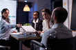© DC Studio - Workaholic disabled leader in wheelchair discussing management statistics solving company graphs. Diverse multi-ethnic businesspeople overworking in meeting room in evening