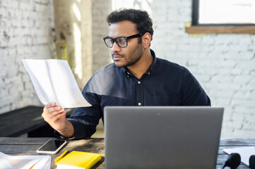 Wall Mural - Executive mixed-race male office employee doing paperwork sitting at the desk with laptop in office, focused and serious hindu man in smart casual looking through sheets of documents, preparing report