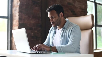 Wall Mural - Indian developer guy is working with a laptop sitting at the desk indoors. A young multiracial guy in smart casual wear is typing on the keyboard, answering email, communicates with the customer