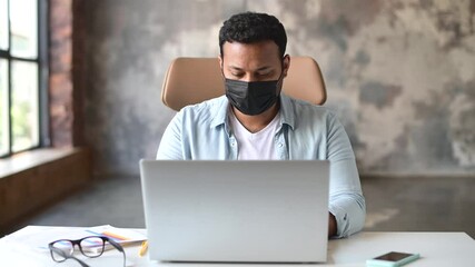 Wall Mural - Confident indian young freelancer man sits at the desk wearing protective mask against coronavirus and flu using laptop computer in the home office, working remotely with face covered