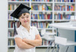 Happy girl with Down Syndrome wearing a graduation cap stands at a library. Empty space for text. Education for disabled children concept