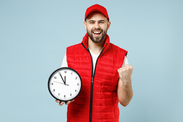 Wall Mural - Professional happy delivery guy employee man in red cap white T-shirt vest uniform workwear work as dealer courier hold clock isolated on pastel blue color background studio portrait. Service concept.