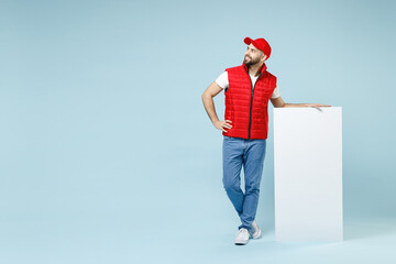 Wall Mural - Full size body length delivery guy employee man in red cap white T-shirt vest uniform work as courier hold big blank poster promo area isolated on pastel blue color background studio. Service concept.