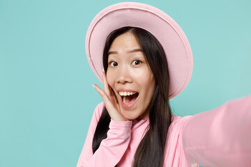 Close up shocked young brunette asian woman 20s wears pink clothes hat do selfie shot on mobile phone keeping mouth wide open looking surprised isolated on pastel blue color background studio portrait