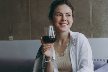 Young smiling happy wistful woman 20s in white clothes plaid sitting on soft grey sofa indoors apartment hold glass drink red wine looking aside Rest on weekends leisure quarantine stay home concept.