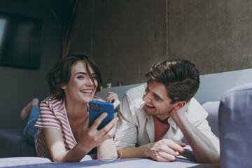 Full body laughing young couple two friends woman man in casual clothes wired earphones lying sofa using mobile cell phone listen to music rest indoors at home flat together People lifestyle concept.