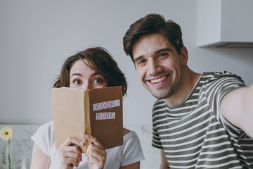 Close up young couple two woman man 20s in casual clothes cover mouth with recipe book do selfie mobile cell phone cook food learn new recipe in light kitchen at home Healthy diet lifestyle concept