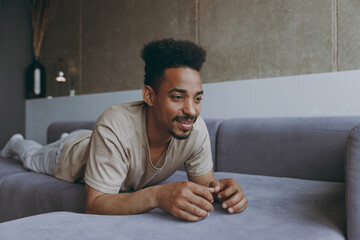 Wall Mural - Pensive smiling wistful brunette young african american man 20s wearing casual beige t-shirt sweatpants lay down on grey sofa indoors apartment resting on weekends staying at home during quarantine.