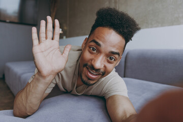 Wall Mural - Close up full length young african american man in t-shirt sweatpants lay down on grey sofa waving hand talk by video call indoors partment do selfie shot on mobile phone rest on weekend stay at home