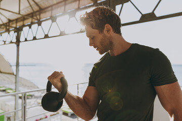 Young serious strong sporty athletic toned fit sportsman man 20s wear sports clothes warm up training work out lifts weight kettlebell at outdoor gym sea beach outdoor seaside in summer day morning.