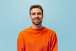 Positive young guy with red beard in bright orange sweatshirt looking into camera and smiling on isolated blue background..