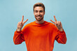 © Look! - Funny trendy man with red beard and brown hair in bright orange outfit smiling and showing peace signs on isolated blue backdrop..