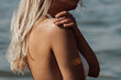 Cropped shot of a young tanned slim blonde woman with a gold boho sticker on her arm touches her shoulder and scrubs the skin with sand on a background of the sea. Close-up. Hot sunny summer day